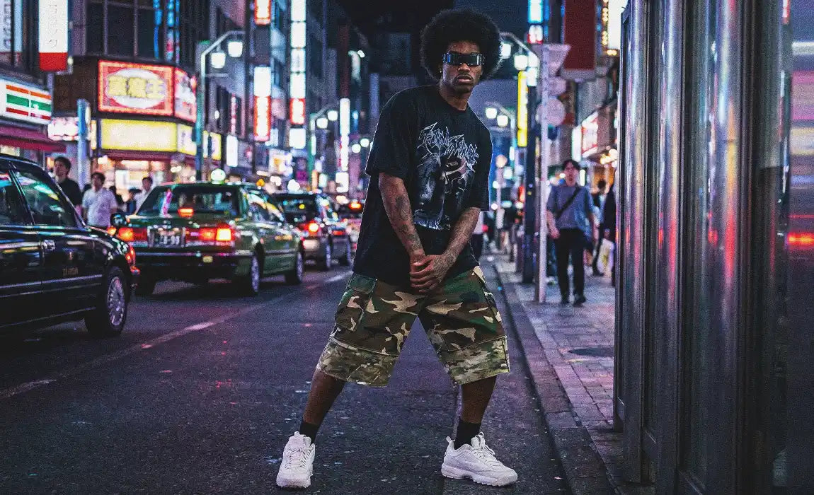 Model in camo cargo jorts and black graphic tee standing on a neon-lit Tokyo street at night with futuristic shield sunglasses and white chunky sneakers