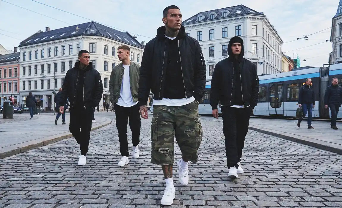 Group of four men walking down a European cobblestone street, center figure wearing distressed camo jorts with a black bomber jacket and white sneakers