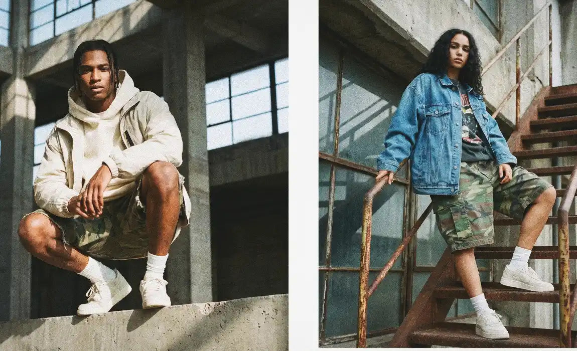 Side-by-side portrait of a male model crouching in ripped camo jorts with a cream hoodie and a female model on rusted industrial stairs wearing camo jorts with an oversized denim jacket