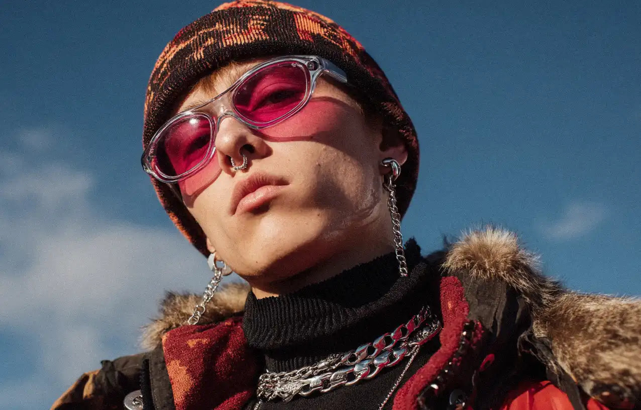 Close-up of a model in pink-tinted sunglasses, layered silver chains, and a beanie, exuding edgy winter street style.