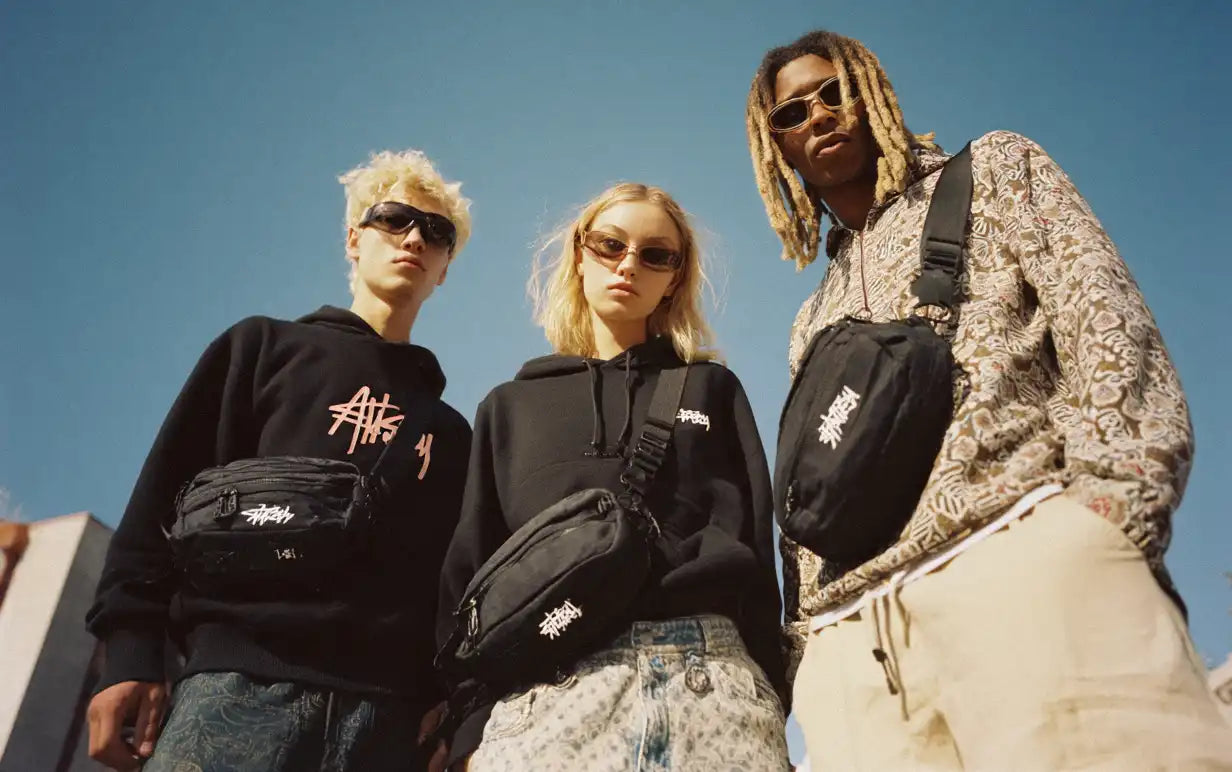 Three young models in streetwear hoodies and sunglasses, all wearing black crossbody bags, under a bright blue sky.