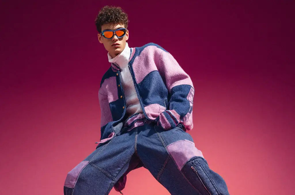 Model in a color-blocked purple and pink denim set with orange reflective sunglasses, posing against a gradient magenta backdrop.