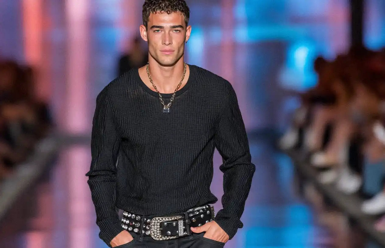 A male model in a black top and jeans is accessorized with a black rhinestone belt.