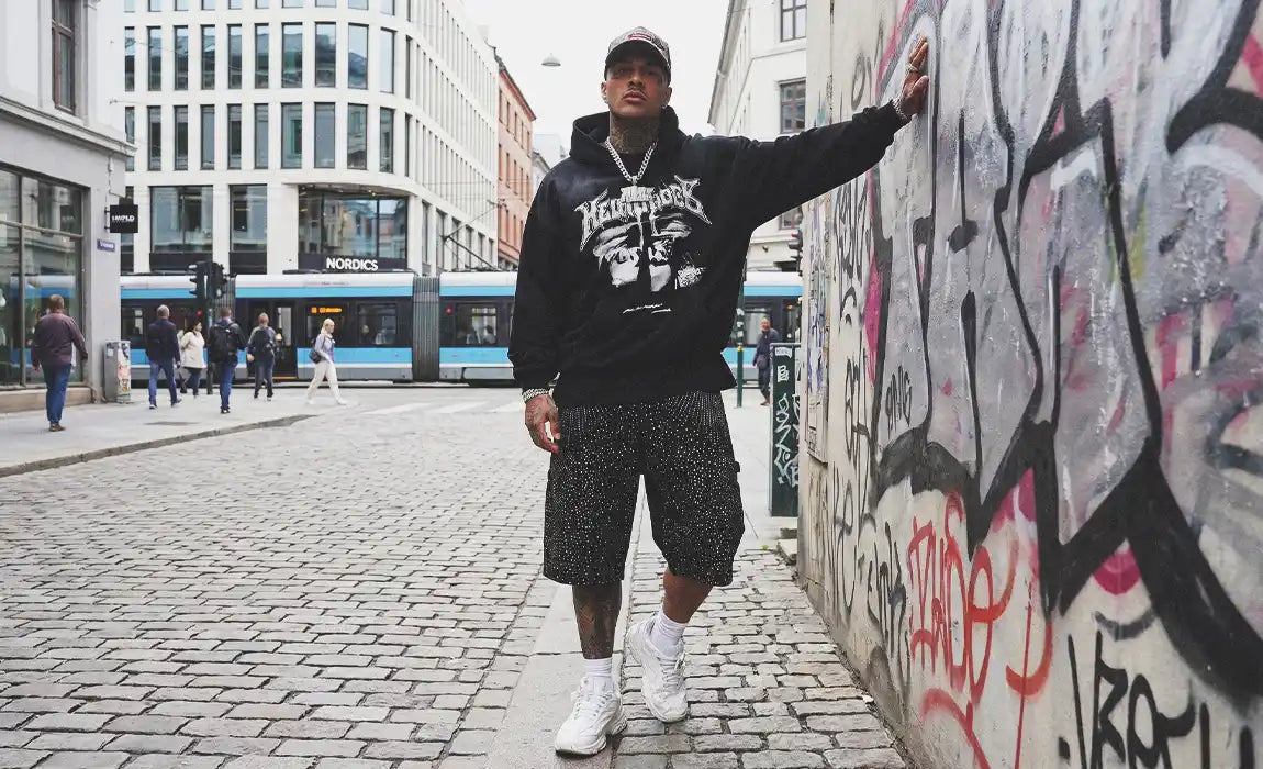 Streetwear model leaning against a graffiti wall wearing baggy black Rhinestone Jorts with an all-over speckled pattern.