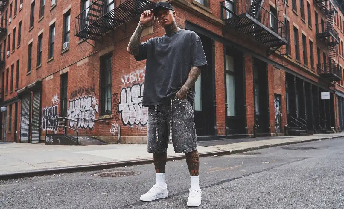 A man in an urban setting wearing dark wash Rhinestone Jorts featuring sparkling dangling straps.
